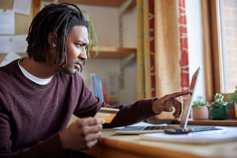 Male University Or College Student With Credit Card Looking At Laptop Worried About Debt At Desk In Room.