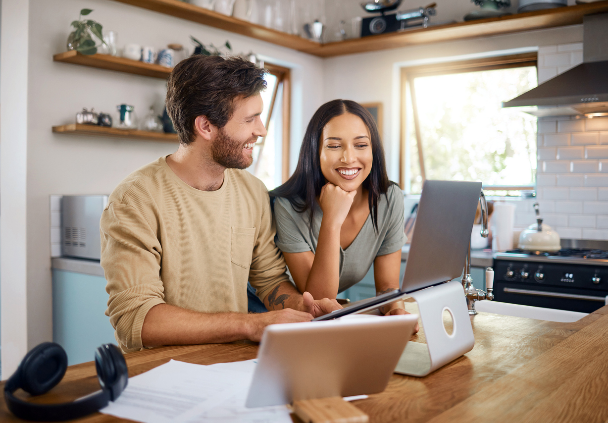 Happy young caucasian man working on laptop while his wife stands next to him looking at the screen.