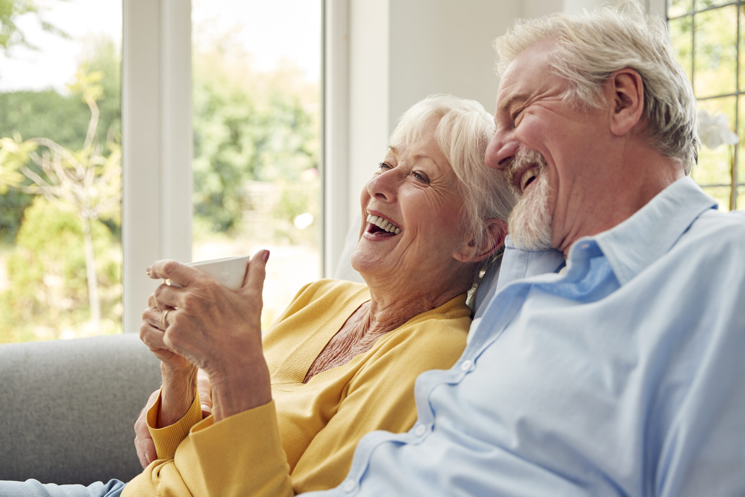 Retired Senior Couple Sitting On Sofa At Home Drinking Coffee And Watching TV Together